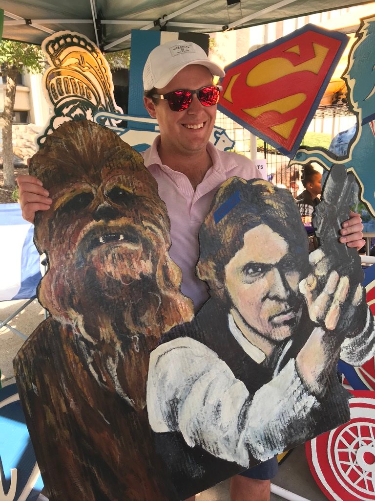 Hand-cut and hand-painted Star Wars portraits of Chewbacca and Han Solo holding a blaster, held by a smiling customer at an outdoor art fair