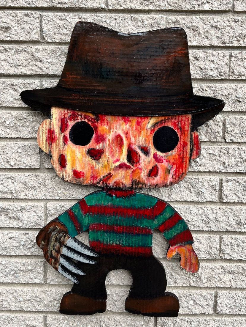 Hand-cut and hand-painted Funko Pop style Freddy Krueger in a brown fedora, red and green striped sweater and a bladed glove, on a brick wall