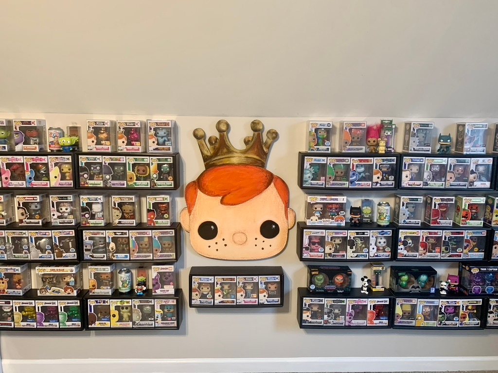 Hand-cut and hand-painted Freddy Funko, a freckled boy with orange hair and a gold crown, hanging between two walls of Funko Pop display boxes