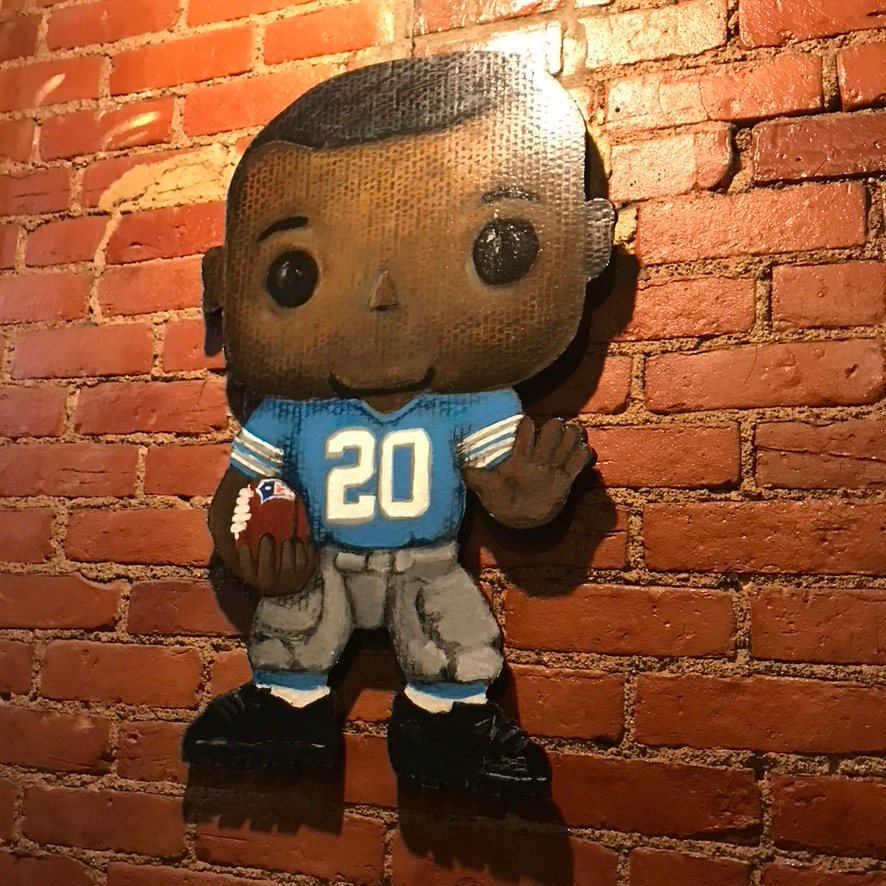 Hand-cut and hand-painted Funko Pop style Detroit Lions running back, number 20, in a blue jersey holding a football, on a brick wall