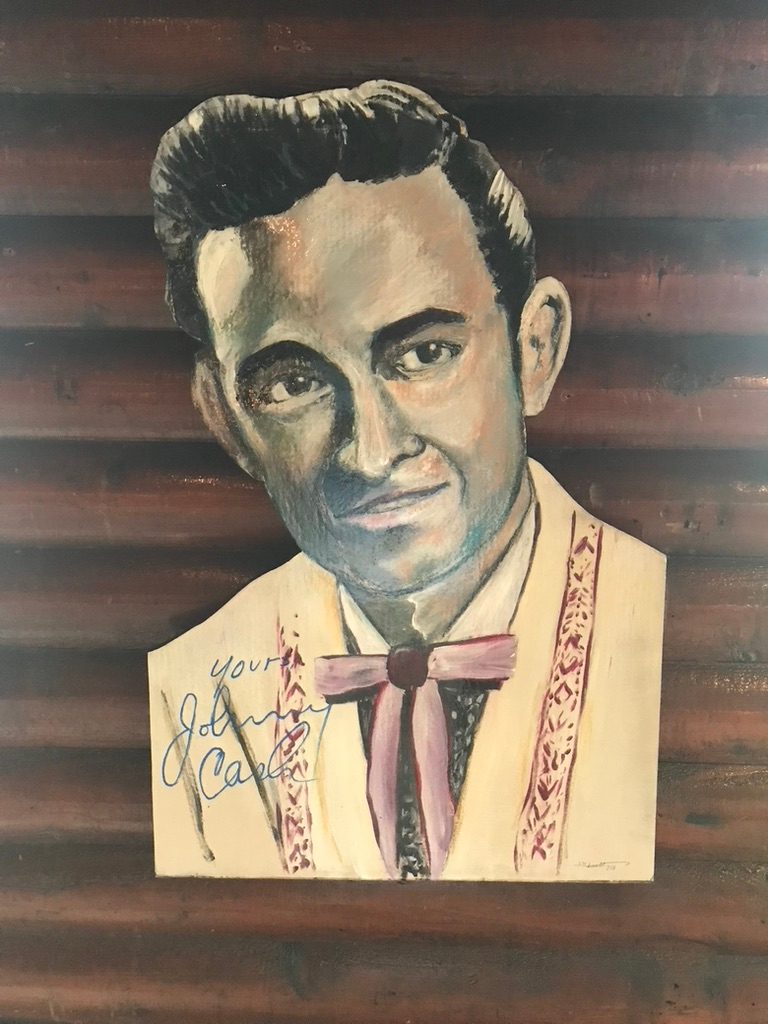 Hand-cut and hand-painted portrait of Johnny Cash in a cream western jacket and pink bolo tie, with a painted signature, on a wood wall