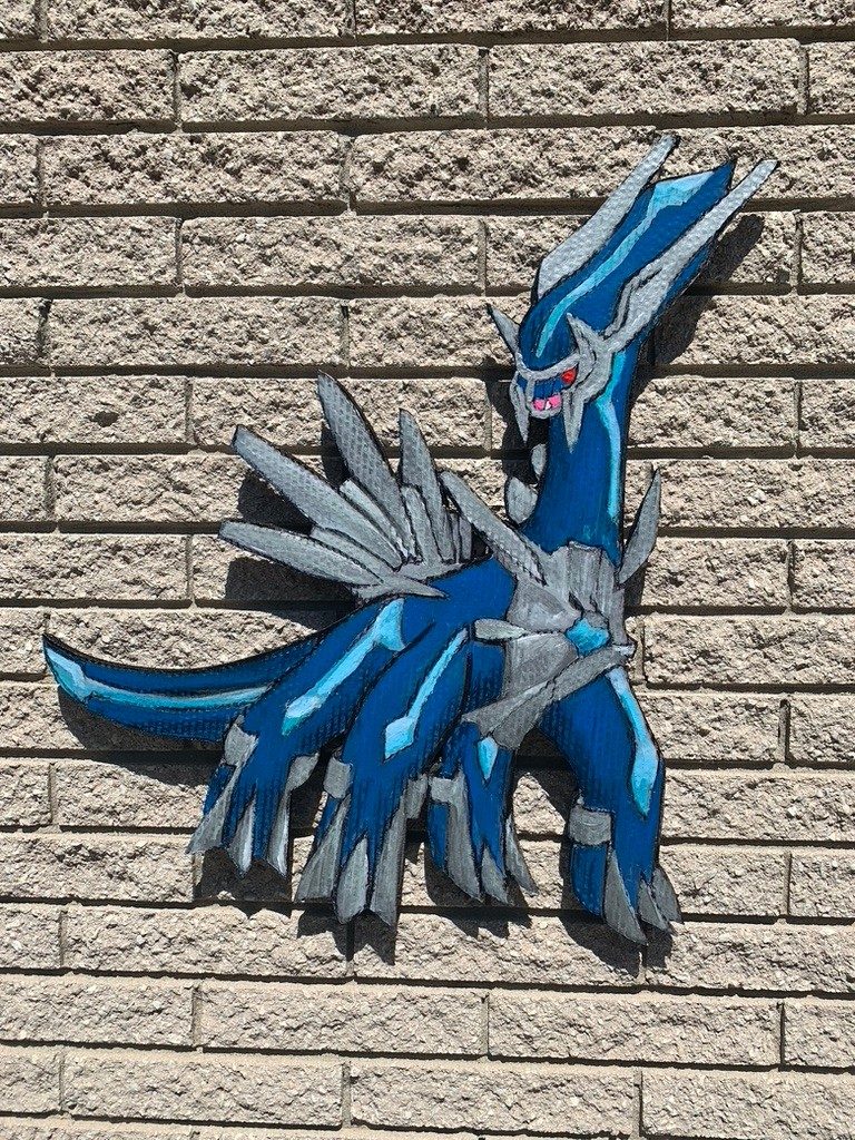 Hand-cut and hand-painted Dialga Pokémon cardboard cutout in blue and silver on a brick wall