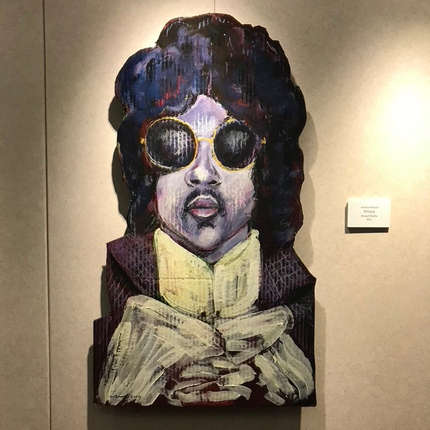 Hand-cut and hand-painted mixed media portrait of Prince in round gold-rimmed sunglasses and a ruffled collar, on a gallery wall