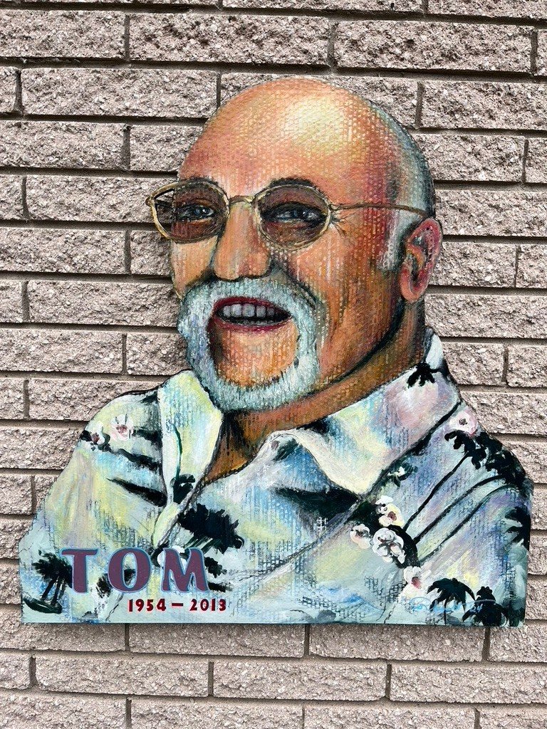 Hand-cut and hand-painted memorial portrait of a smiling man with a gray beard and round glasses in a tropical shirt, lettered Tom 1954 to 2013, on a brick wall
